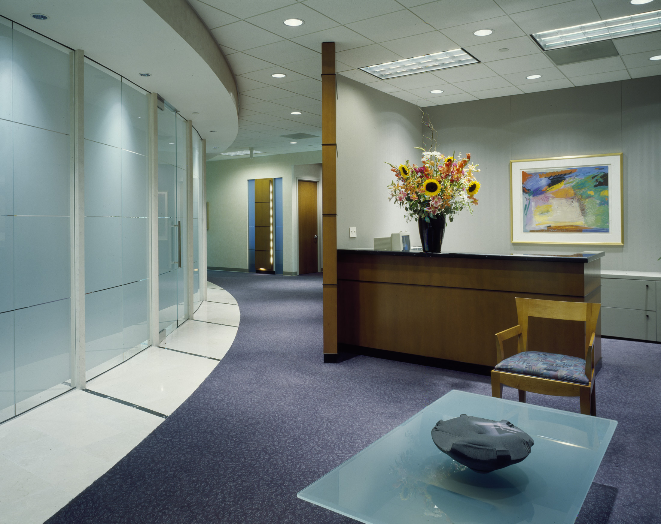 a brightly lit reception room from the 1980s is shown with a rounded glass wall to the left and a glass coffee table in the foreground over purple carpet, a wood desk is in the back with a large colorful floral bouquet 