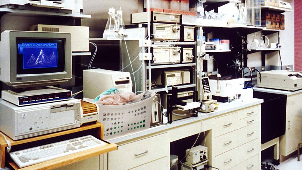 A laboratory is seen with an old computer showing a blue mountain range on it and next to the computer is a basket of laundry