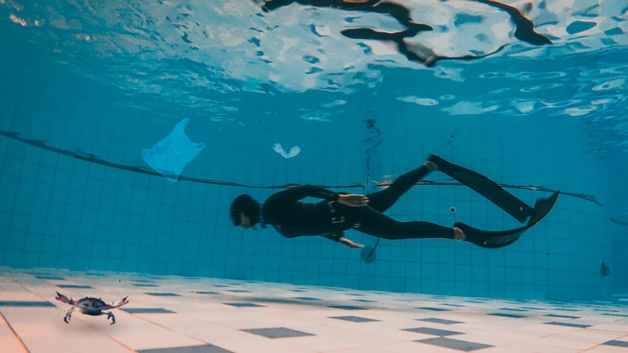 A person scuba diving in a swimming pool with some loose garbage and a small animated crab at the bottom of the pool with their claws up