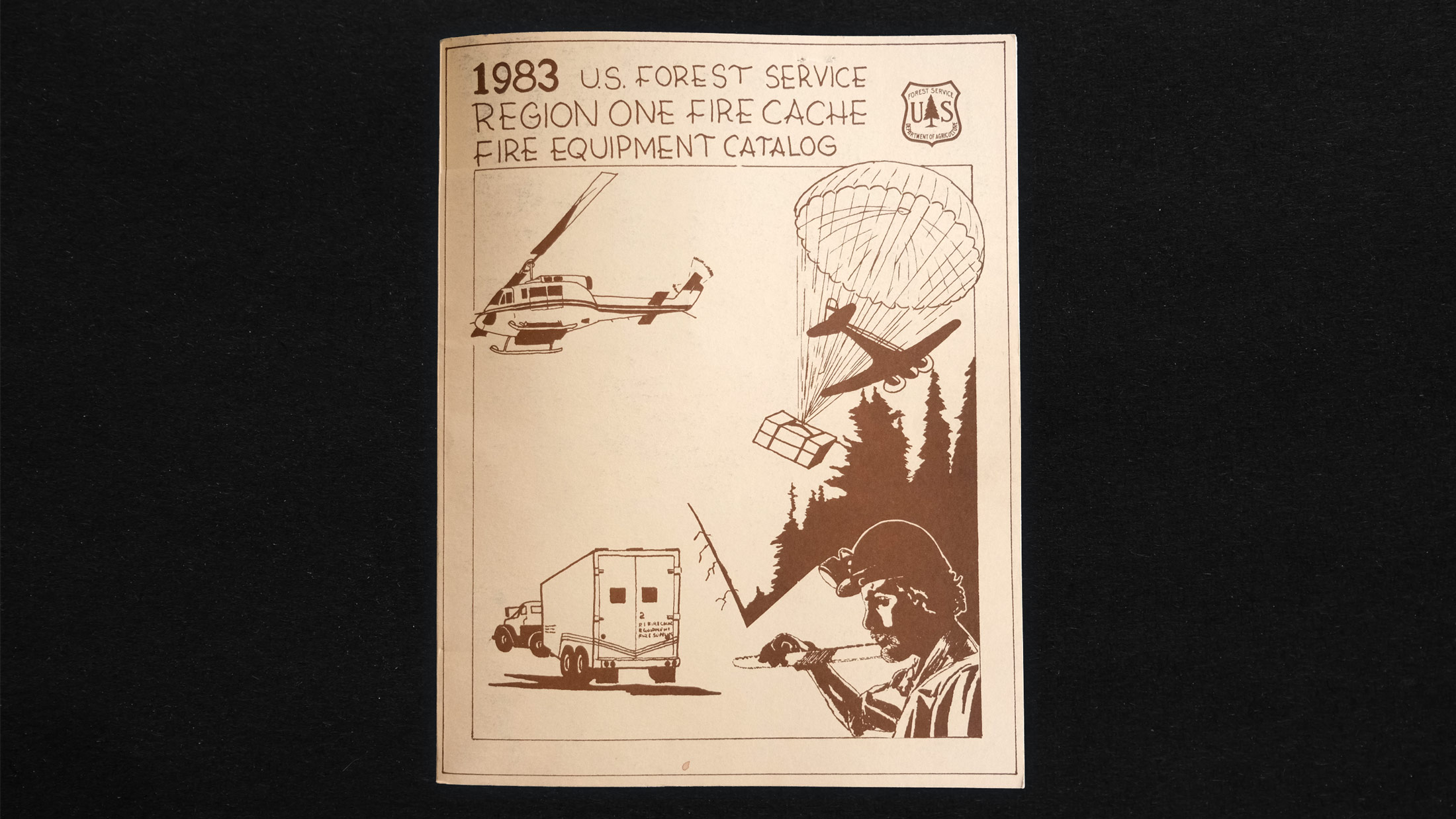 A scene of a helicopter, airplane, truck, and lumberjack near a forest on the cover of US Forest Service Region One Fire Cache Fire Equipment Catalog (1983) by the US Forest Service