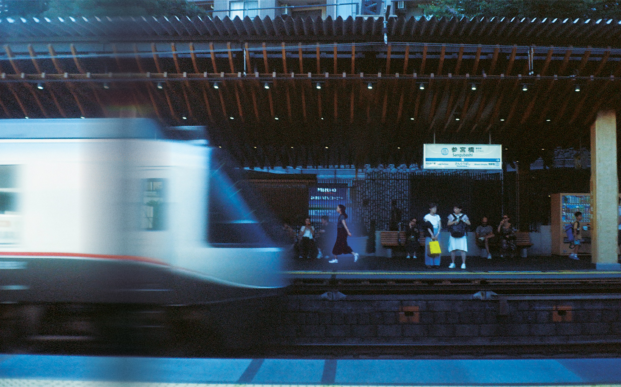 A photo of a train arriving at a station platform in tokyo as a woman and others wait for it to stop